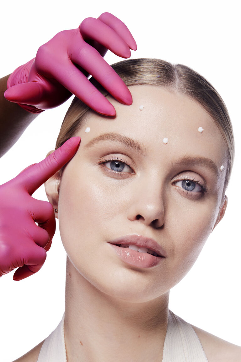 Woman with dots of Mary Kay Dynamic Wrinkle Limiter on her forehead. A hand with pink gloves is touching her forehead to give scientific effect.