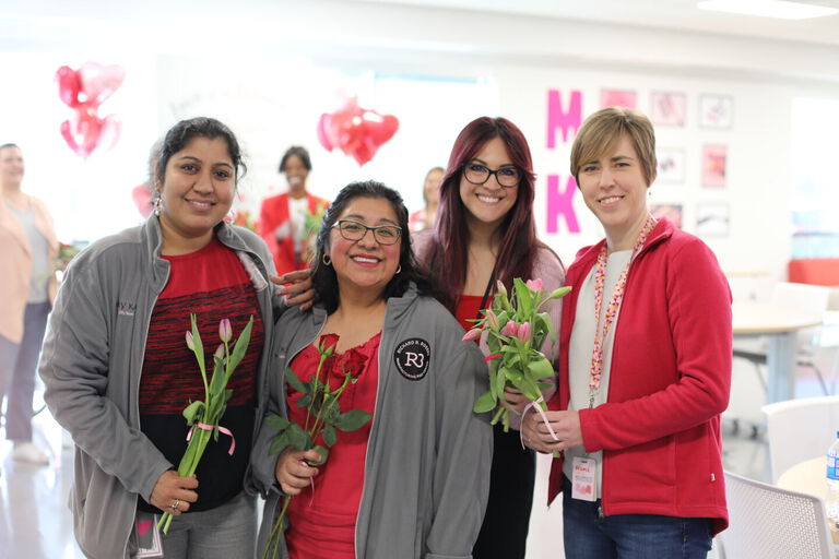 Grupo de colaboradores de Mary Kay sosteniendo flores durante un evento en interiores, con globos en forma de coraz&oacute;n al fondo.