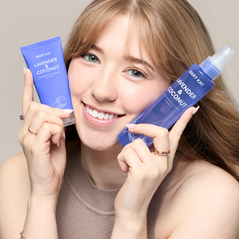 A white model with blue eyes holds the lavender and coconut hand cream and body spray by her face while smiling.&nbsp;&nbsp;&nbsp;