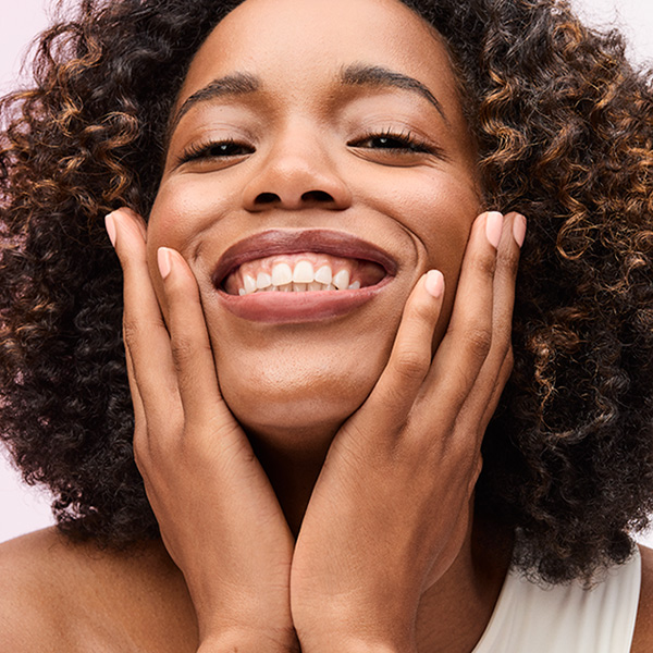 A woman with radiant, well-cared-for skin rests her face in her hands and smiles at the camera.
